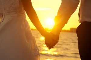 Young adult male groom and female bride holding hands on beach at sunset.